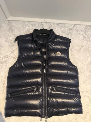 Mörkblå Moncler Gui väst - Tjena, säljer nu en mycket populär Moncler Gui väst i nyskick, den är använd fåtal gånger och har inga defekter. De följer med en liten bag där man kan packa ner den (syns på bild 3). Den är storlek 3. Och såklart äkta! Har du någon fundering är de bara att skriva, ge gärna prisförslag!!