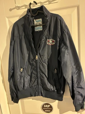 Mörkblå bomberjacka Biaggo Jeans - Snygg mörkblå bomberjacka från Bugatchi Jeans med broderade detaljer på bröstet och ryggen. Jackan har dragkedja framtill, två fickor med dragkedja och ribbade muddar vid ärmslut och midja. Patch med siffran 84 på ena ärmen ger extra stil.
