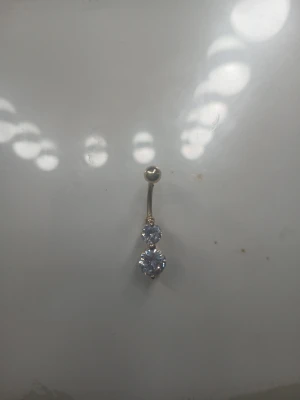 Smyckad navelpiercing med stenar - Snygg navelpiercing i silverfärgad metall med två glittrande, runda klara stenar. Klassisk böjd stav och kula som skruvas av för enkel användning. Perfekt för dig som vill ha något stilrent men ändå glittrigt till din piercing.