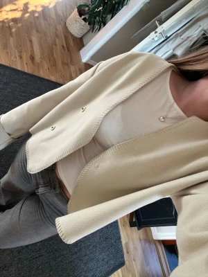 Beige croppad kofta med knappar från Gina Tricot  - Stilren beige kofta/tunnare jacka med croppad passform och vida ärmar. Koftan har dubbla knappar framtill och är tillverkad i ett mjukt, tunt material som ger en avslappnad look. Perfekt att slänga över en t-shirt för en enkel men snygg outfit. Någon liten fläck (se sista bild)