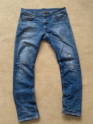 Rare Blå slitna jeans med hål från nudie St 34,34 - Ett par blå jeans i klassisk modell med slitningar och hål på höger ben. Jeansen har fem fickor, normal midja och raka ben. Materialet är denim i bomull och de har en avslappnad, trendig look. De har gått sönder i skrevet men de är åtgärdat. Storlek 34-34. Hör av er vid minsta fundering 💫