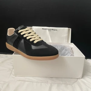 Maison Martin Margiela  - Maison Margiela sneakers i svart läder och mocka med beigea skosnören och klassisk gummisula. Snygg retrostil med låg profil och diskreta detaljer. Perfekta för dig som gillar stilrena och tidlösa sneakers. de kommer med förpackning och är köpta på en second hand hemsida men är helt nya, jag säljer de pga att de inte passar mig längre. 