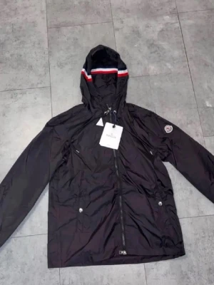 Svart vindjacka med huva Moncler - Snygg svart vindjacka från Moncler med huva och dragkedja. Jackan har vita och röda detaljer på huvan, två fickor med knappar och Moncler-logga på ärmen. Tillverkad i lätt, glansigt material som står emot vind. Perfekt för dig som vill ha en stilren och sportig look.