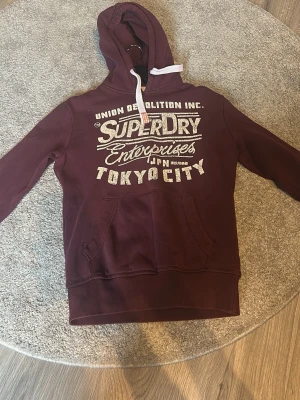 Vinröd hoodie från Superdry - Snygg vinröd hoodie från Superdry med stor vit logga och text på bröstet. Klassisk känguruficka framtill, huva med vita dragsnören och ribbade muddar. Perfekt för en avslappnad look.