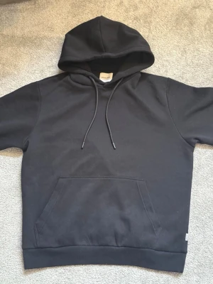 Svart hoodie från Jack & Jones - ANVÄND 3 GÅNGER! Säljer en svart hoodie från Jack & Jones med klassisk huva och dragsnören. Hoodien har en stor magficka framtill och ribbade muddar vid ärmslut och nederkant. Tillverkad i mjukt bomullsmaterial som är skönt att bära.