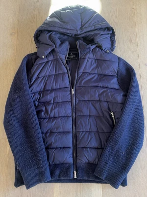 Mörkblå Cardigan Jacka - Snygg mörkblå pufferjacka med stickade ärmar och avtagbar huva. Jackan har dragkedja framtill och två sidofickor med dragkedja. Materialmix med quiltad front och rygg i pufferstil samt stickade partier på ärmar och sidor. Perfekt för kyliga dagar.