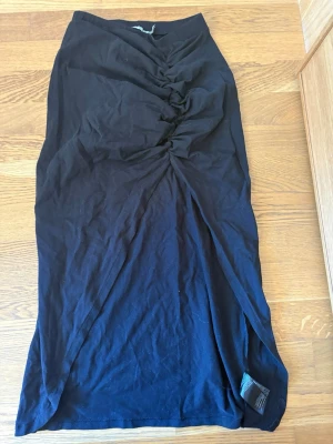 Svart långkjol med slits från NA-KD - Svart långkjol från NA-KD i mjukt material. Kjolen har en snygg drapering framtill och en hög slits på sidan som ger en trendig look. Perfekt att styla med en crop top eller oversized tröja.