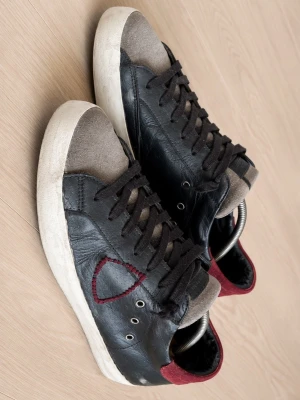 Philippe Model svarta sneakers strl 41 - Snygga svarta Philippe Model sneakers med grå mockadetaljer och vinröd häl. Skorna har snörning, rund tå och vit platt sula. Märkets logga syns på sulan och insidan. Materialmix av skinn och mocka ger en cool kontrast. Perfekt för dig som gillar streetstyle.