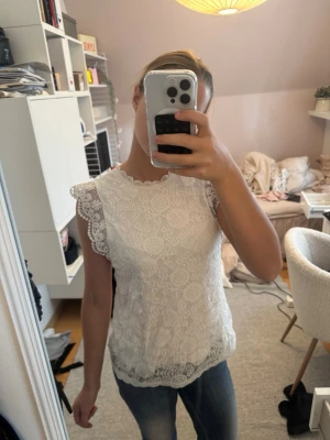 Vit spetsblus med korta ärmar - Säljer en superfin vit blus i spets med detaljerade mönster och vågig kant vid halsen. Blusen har korta spetsärmar och är lätt transparent. Perfekt att styla med jeans eller kjol för en fräsch look.