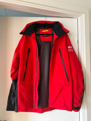 Röd seglarjacka sailracing - Snygg röd vindjacka med svarta detaljer och dragkedjor framtill. Jackan har huva, två stora bröstfickor med dragkedja och ett tryckt vitt märke på ärmen. Materialet är tunt och vindtätt, perfekt för blåsiga dagar. Sportig design med svart insida och justerbara ärmslut.