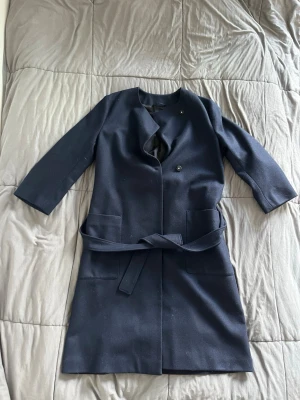 Mörkblå kappa från Filippa K - Stilren mörkblå kappa från Filippa K med knytband i midjan och stora fickor framtill. Kappan har trekvartsärmar och en minimalistisk design utan synlig krage. Perfekt för dig som gillar klassisk och enkel stil.