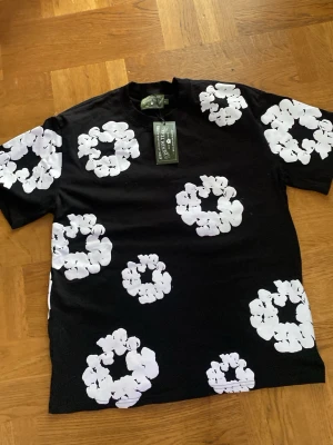 Svart t-shirt med vita blommor Denim Tears - Säljer en svart t-shirt från Denim Tears med stora vita blommönster över hela plagget. T-shirten har rund hals och korta ärmar. Perfekt för dig som vill sticka ut med en unik och trendig look.