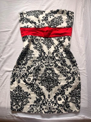Vintage strapless klänning med röd midja - Säljer en snygg vintage axelbandslös klänning från ADK i storlek 44. Klänningen är vit med svart barockinspirerat mönster och har ett brett rött band i midjan. Perfekt för dig som vill sticka ut med en elegant och unik look.