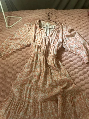 Blommig långklänning från s.Oliver - Långklänning från s.Oliver i ljusa toner med blommigt mönster i vitt och orange. Klänningen har v-ringning, volangdetaljer och trekvartsärm. Perfekt för sommarens alla tillfällen.