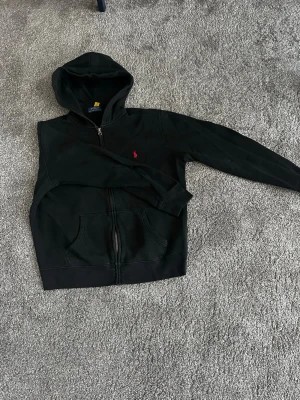 Svart hoodie från Polo Ralph Lauren - Svart hoodie med dragkedja från Polo Ralph Lauren. Klassisk design med huva, kängurufickor och det ikoniska röda broderade logotypen på bröstet. Tillverkad i mjukt bomullsmaterial som är skönt att bära. Perfekt för en avslappnad och stilren look.