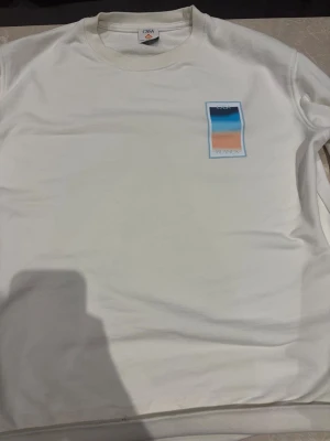 Vit sweatshirt från Casa Blanca - Snygg vit sweatshirt från Casa Blanca med en färgglad grafisk patch på bröstet i blått, orange och beige. Tillverkad i mjuk bomull och har en klassisk rund halsringning. Perfekt för en clean och stilren look.