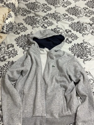 Grå zip hoodie från Lacoste - Snygg grå hoodie med dragkedja från Lacoste. Klassisk krokodil-logga på bröstet och två fickor framtill. Huvan har mörkblått foder och tröjan är gjord i mjukt sweatshirtmaterial.. köptes för 