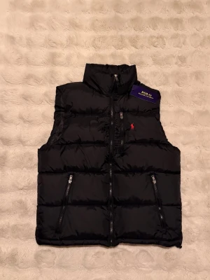 Väst från Polo Ralph Lauren - Snygg svart dunväst från Polo Ralph Lauren med hög krage och två dragkedjefickor framtill. Västen har det klassiska röda Polo-logot broderat på bröstet och en glansig finish. Perfekt för lager på lager och streetwear-vibe.