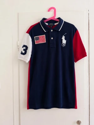 Snygg Polo Ralph Lauren Big Pony piké i marinblått, rött och vitt 🇺🇸 - Klassisk Ralph Lauren-design med stor broderad Big Pony-logga på bröstet, USA-flagga och nummer 3 på ärmen. Riktigt snygg colorblock-design med röd rygg. • Storlek M (10–12) • Polo Ralph Lauren • Big Pony • USA-flagga på bröstet • Broderade detaljer • Marinblå/röd/vit Fint begagnat skick. Se bilder för skick. Skriv gärna om du vill ha mått eller fler bilder!