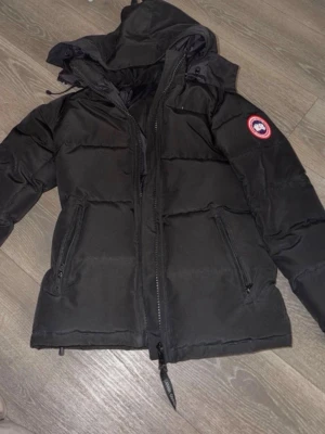 Svart dunjacka  - Snygg svart dunjacka från Canada Goose med huva och dragkedja framtill. Jackan har två dragkedjeförsedda fickor och den klassiska logotypen på ärmen. Perfekt för kalla dagar och riktigt varm tack vare dunfyllningen.