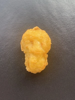 Kycklingnugget med unik form - En gyllene, friterad kycklingnugget med en ovanlig form som påminner om ett huvud eller en skalle. Perfekt för dig som gillar roliga och annorlunda snacks. Nuggeten har en krispig yta och är lagom stor för ett mellanmål.