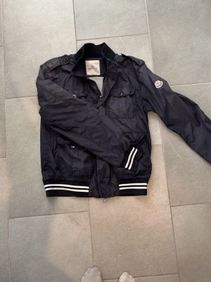 Svart vindjacka från Moncler - 100% äkta Snygg svart vindjacka från Moncler med vita ränder på muddar och midja. Jackan har flera fickor framtill, axelklaffar och Moncler-logga på ärmen. Tillverkad i lätt, glansigt material som passar perfekt till streetwear. Passar 170 små hål vid armarna