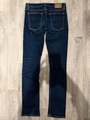 Nudie Jeans Tube Kelly Mörkblå W29/L34 - Slim Fit - Nudie Jeans Tube Kelly ”Rinsed Strikey” mörkblå jeans med slim fit-passform, normalhög midja och smala, avsmalnande ben, storlek W29/L34, midja 38,5 cm, ytterbenslängd 107,5 cm, benöppning 16,5 cm, modellen är 185cm & väger 65kg, i fint skick.