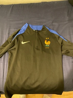 Nike Frankrike träningströja svart/blå säljer byxorna oxå - Snygg svart träningströja från Nike med blå detaljer på axlarna och halv dragkedja. Bröstet pryds av Frankrikes landslagsmärke med tupp och två stjärnor samt Nike-logga. Perfekt för fotbollsträning eller chill. Materialet är troligtvis polyester.