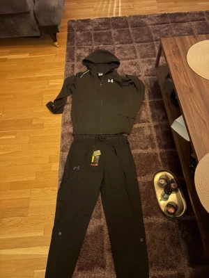 Svart träningsset från under armour det står XL på tagsen men den passar M den är väldigt liten i storlek  - Säljer ett svart träningsset från Under Armour med huva och dragkedja på tröjan samt matchande byxor med snörning i midjan. Setet är gjort i ett lätt och funktionellt syntetmaterial, perfekt för träning. Under Armour-logga finns på både tröja och byxa.