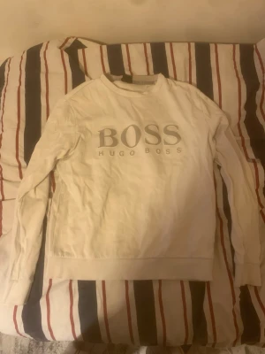 Vit sweatshirt från Hugo Boss - Snygg vit sweatshirt från Hugo Boss med stort reflekterande BOSS-tryck på bröstet. Tröjan har rund halsringning, långa ärmar och ribbade muddar. Perfekt för dig som gillar stilrena och klassiska plagg med en twist.