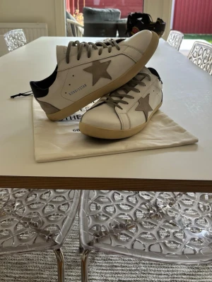Golden Goose sneakers - Snygga vita sneakers från Golden Goose med grå stjärna på sidan, grå skosnören och beige sula. Skorna har svart detalj vid hälen och är tillverkade i läder. Perfekta för dig som gillar en stilren men ändå unik look. Andvända 3 gånger