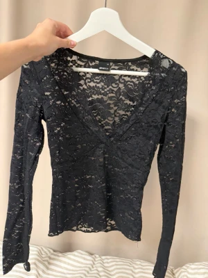Svart långärmad spetsblus - Snygg svart långärmad topp i genombruten spets med djup V-ringning. Figurnära modell som ger en elegant look. Perfekt att styla med ett linne under eller som den är för en trendig outfit.