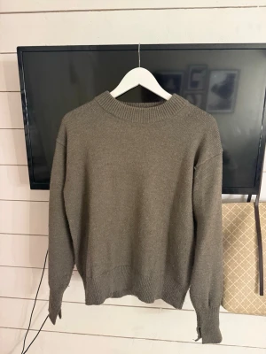 Olivgrön stickad tröja med rund hals - En enkel och stilren olivgrön stickad tröja med rund halsringning och ribbade muddar vid ärmslut och nederkant. Tröjan har en avslappnad passform och är perfekt för lager på lager. Materialet känns mjukt och bekvämt.