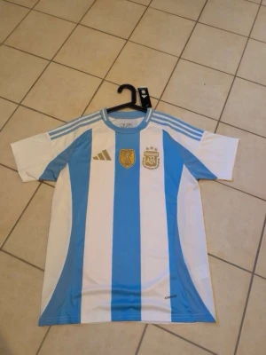 Argentina fotbollströja blåvit Adidas - Säljer en officiell Argentina fotbollströja från Adidas i blått och vitt med klassiska ränder. Tröjan har guldfärgade detaljer, AFA-emblem och FIFA-märke på bröstet samt korta ärmar och rund hals. Perfekt för dig som älskar fotboll och vill stötta ditt lag.