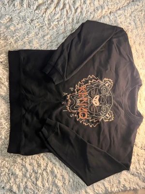 Svart Kenzo sweatshirt med tigertryck - Svart sweatshirt från Kenzo med ikoniskt broderat tigerhuvud och texten 'KENZO PARIS' i orange och beige. Tröjan har rund hals, långa ärmar och är gjord i mjukt bomullsmaterial. Perfekt för dig som vill sticka ut med en streetstyle-look.