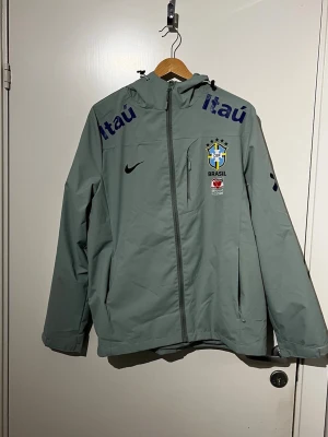 Grå Brasil Nike vindjacka med huva - Snygg grå vindjacka från Nike med Brasilien CBF-logga och sponsorer tryckta på bröstet. Jackan har huva med dragsko, hel dragkedja framtill och flera fickor. Perfekt för dig som gillar sportig stil och vill representera Brasilien.
