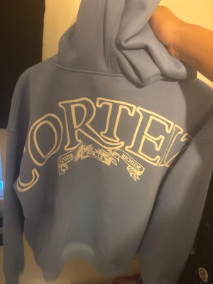 Blå Corteiz double zip hoodie  - Säljer en ljusblå hoodie från Corteiz med stor vit logga och text på bröstet samt mindre tryck på framsidan. Hoodien har dragkedja, fickor framtill och en klassisk huva. Perfekt för dig som gillar streetwear och vill sticka ut.