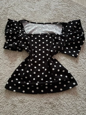 Svart prickig topp med puffärm - Snygg svart topp med vita prickar och puffiga korta ärmar. Fyrkantig ringning ger en trendig look och materialet känns mjukt och följsamt. Perfekt för dig som vill sticka ut lite extra med ett klassiskt mönster.