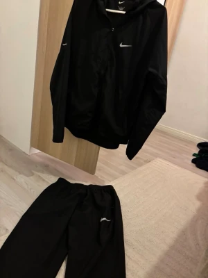 Svart Nike Miller - Säljer ett svart träningsset från Nike med jacka och byxor. Jackan har dragkedja, huva och Nike-logga på bröstet. Byxorna har resår i midjan och Nike-logga på benet. Perfekt för träning eller uppvärmning.