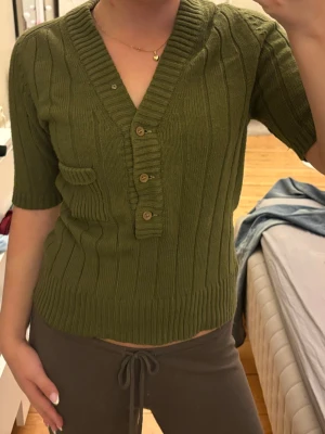 Olivgrön stickad topp med knappar - Snygg olivgrön stickad topp med ribbad struktur, djup v-ringning och fyra träknappar framtill. Kort ärm och liten dekorficka på ena sidan. Perfekt till jeans eller kjol för en trendig look💗🪷 OBS! Står att det är storlek 42 men sitter som S!