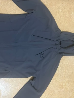 Grå vindjacka från Under Armour - Snygg grå vindjacka från Under Armour. Jackan är oanvänd eftersom jag köpte fel storlek och den är stor på mig. Jackan har hel dragkedja framtill och diskret logga på bröstet. Tillverkad i ett lätt och tåligt material som står emot vind. Perfekt för dig som vill ha en stilren och funktionell jacka. 