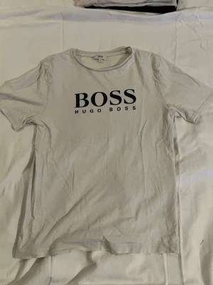 Vit kortärmad t-shirt från Hugo Boss - Snygg vit t-shirt från Hugo Boss med klassisk rund hals och stor svart logga på bröstet. Tillverkad i mjuk bomull för en bekväm känsla. Perfekt till jeans eller shorts för en clean och stilren look.