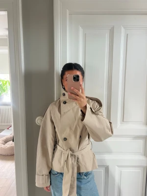 Kort beige trenchcoat med bälte - Stilren beige trenchcoat med dubbelknäppning, höga krage knytskärp i midjan. Jackan har vida ärmar med knappdetaljer vid ärmsluten och axelklaffar. 