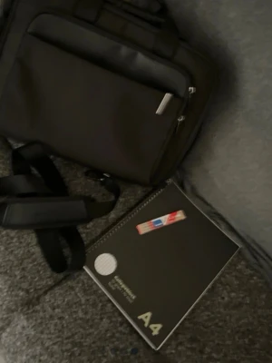 Svart datorväska med axelrem - Snygg svart datorväska i rektangulär form med praktiskt ytterfack och justerbar axelrem. Perfekt för att bära laptop och tillbehör till skolan eller jobbet. Väskan har handtag upptill och är tillverkad i slitstarkt material.