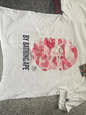 Vit BAPE t-shirt med rosa camomönster - Säljer en vit t-shirt från BAPE med stort rosa camomönstrat apansikte på bröstet och texten 'BY BATHINGAPE' i svart. Klassisk passform och rund hals. Perfekt för dig som gillar streetwear och vill sticka ut. Lite damged