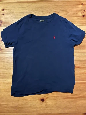 Polo Ralph Lauren  - Klassisk mörkblå t-shirt från Polo Ralph Lauren med röd broderad logga på bröstet. T-shirten har rund halsringning och korta ärmar. Tillverkad i mjuk bomull för en skön känsla och enkel stil.