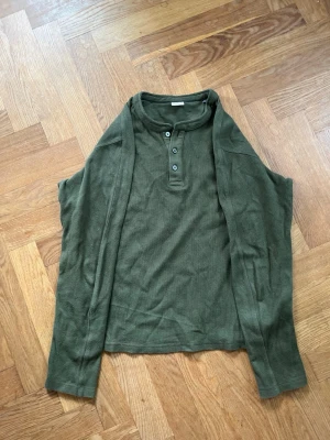 Olivgrön långärmad henleytröja - Använd endast en gån. En olivgrön Jack and Jones långärmad henleytröja med tre knappar vid halsen. Tröjan har en avslappnad passform och är tillverkad i ett mjukt, lite tjockare tyg. 