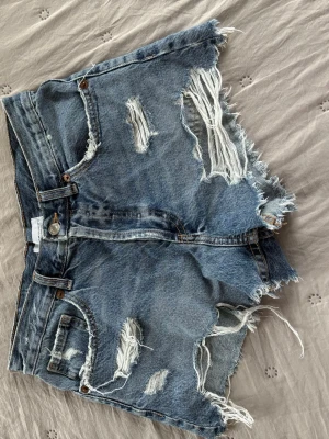 Blå slitna jeansshorts med fransar - Säljer ett par blå jeansshorts med slitna detaljer. Shortsen har medelhög midja, fem fickor och fransar längs bensluten.