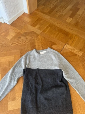 Gråmelerad stickad tröja - Säljer en stilren stickad tröja i två nyanser av grått. Tröjan har rund halsringning, långa ärmar och ribbade muddar vid ärmslut och nederkant. Perfekt för lager på lager och passar till det mesta i garderoben. Nytt skick! Storlek medium!