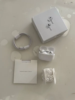 AirPods Pro  - Hej Säljer ett par vita trådlösa  hörlurar med laddningsetui, extra silikonpluggar och laddkabel. Hörlurarna har en stilren och modern design med rundade former och touchkontroller på sidan. Perfekt för musik, poddar och samtal. 